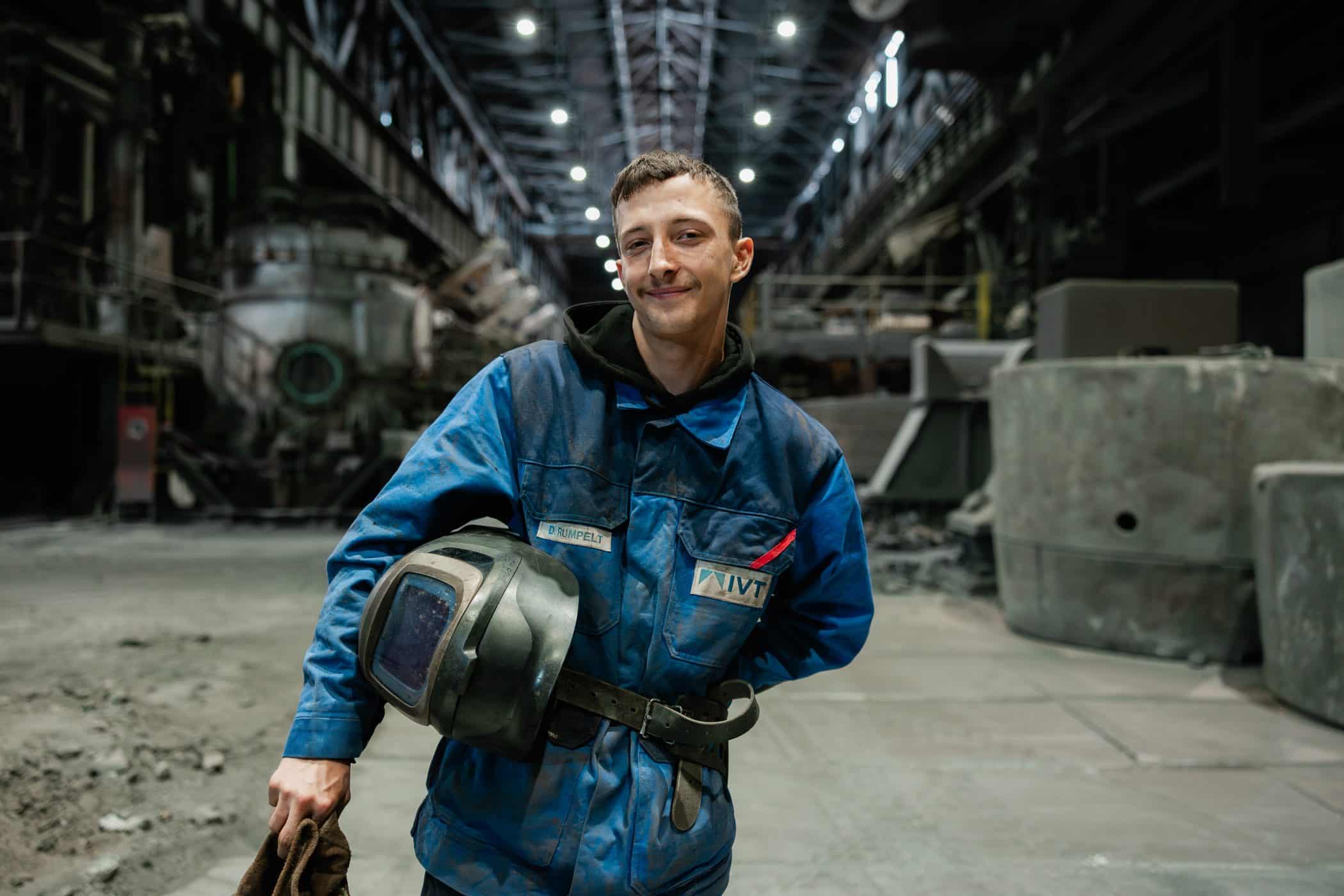 Junger Mitarbeiter in Arbeitskleidung haelt Schweisshelm in Werkhalle bei IVT in Oberhausen