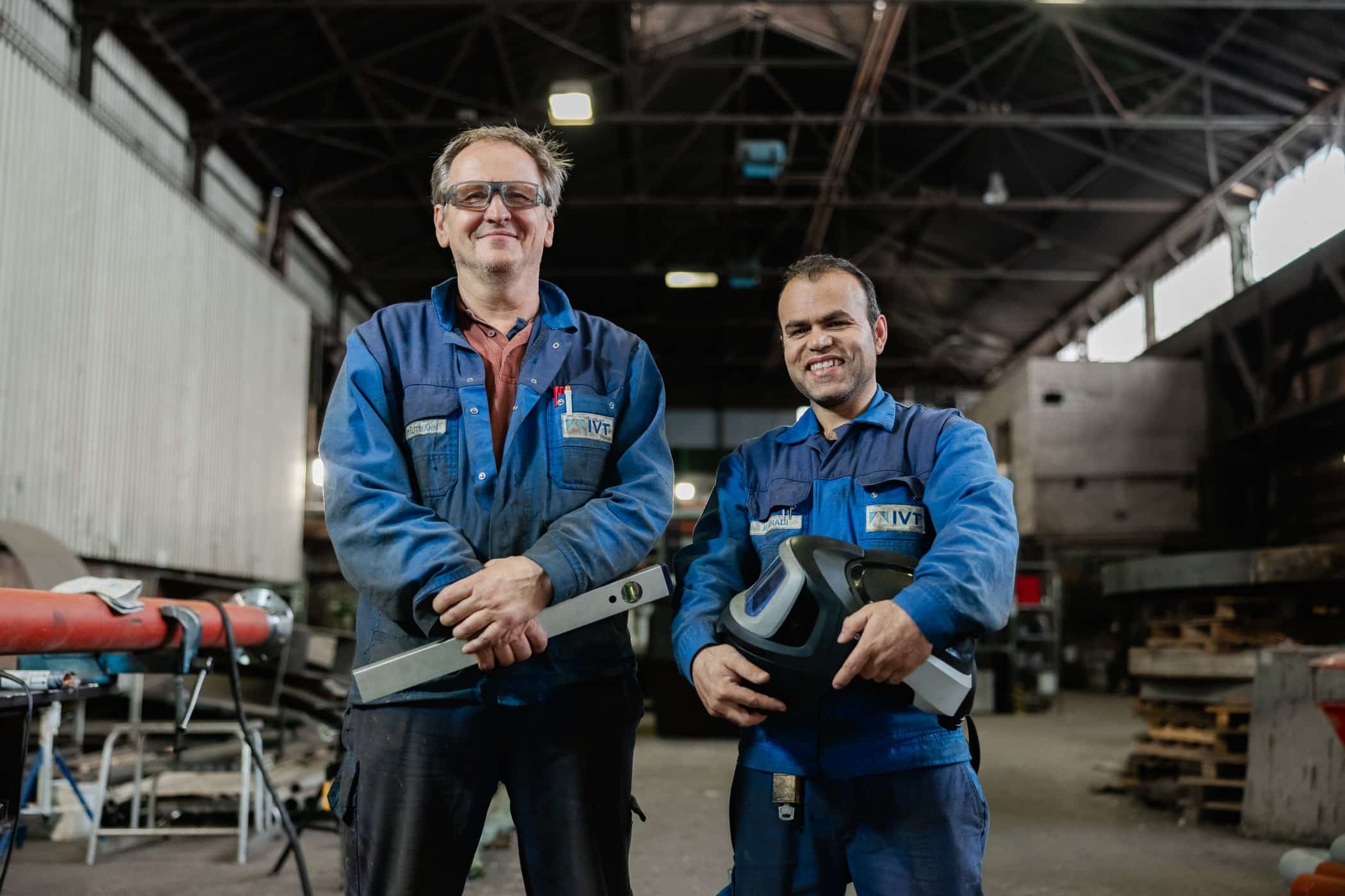 Zwei Mitarbeiter stehen in Werkhalle und laecheln in die Kamera