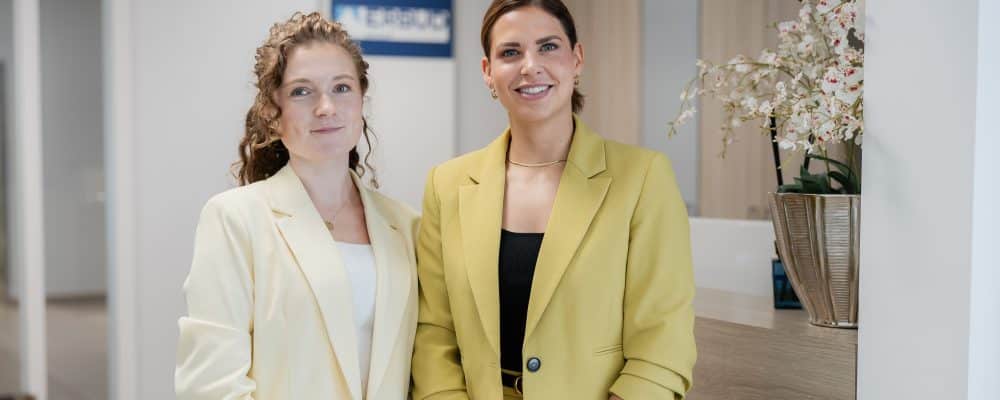 Zwei Mitarbeiterinnen stehen am Empfang und laecheln in die Kamera