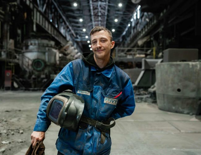 Junger Mitarbeiter haelt Schweisshelm in grosser Produktionshalle